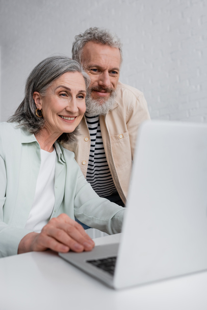 Positive freelancer using laptop near middle aged husband at home  - Foto, Imagem