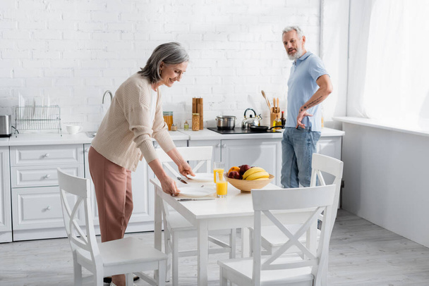 Smiling woman putting plates on table near orange juice, fruits and husband in kitchen  - Foto, Imagem