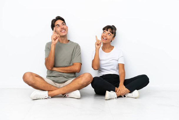 Young mixed race couple sitting on the floor isolated on white background pointing a great idea and looking up - Photo, Image