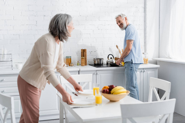 Positive mature man cooking and looking at wife with plates in kitchen  - Foto, afbeelding