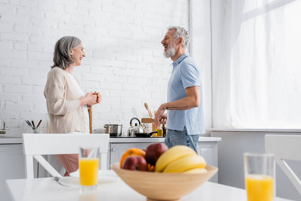 Side view of cheerful mature couple talking near stove in kitchen  - Foto, Bild