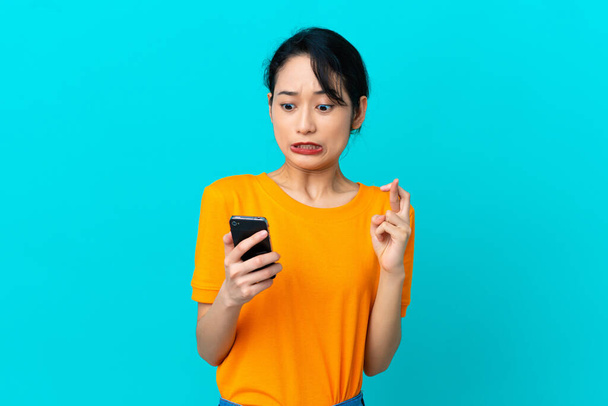 Young Vietnamese woman isolated on blue background using mobile phone with fingers crossing - Foto, Imagem