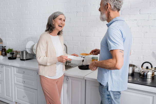 Positive woman holding plate near husband with frying pan and pancake in kitchen  - Photo, Image
