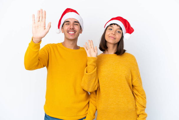 Mladý smíšený závod pár slaví Vánoce izolované na bílém pozadí salutování s rukou s veselým výrazem - Fotografie, Obrázek