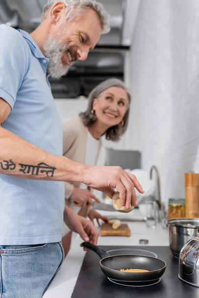Smiling man holding spice while cooking pancake near blurred wife in kitchen  - Photo, Image