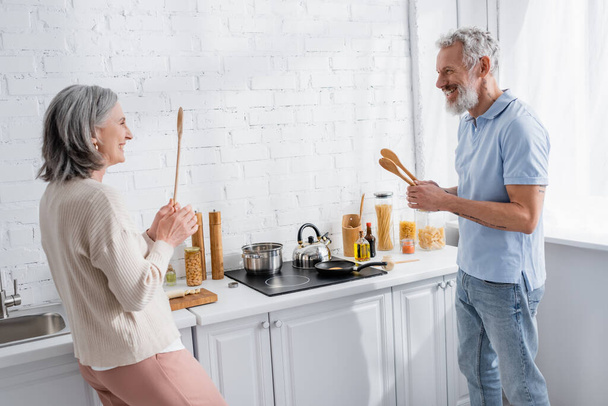 Happy mature couple holding spoons near stove in kitchen  - Фото, зображення