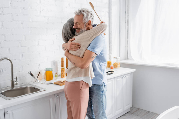 Smiling man hugging wife with spoon in kitchen  - Fotó, kép