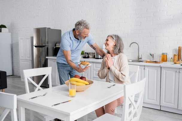 Mężczyzna trzymając talerz z naleśnikami podczas uśmiechniętej żony pokazując proszę gest w kuchni  - Zdjęcie, obraz