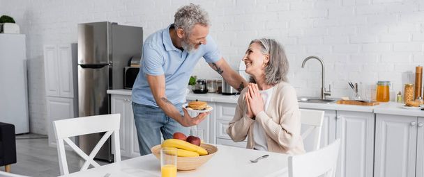 Smiling woman showing please gesture near mature husband with pancakes in kitchen, banner  - Photo, Image