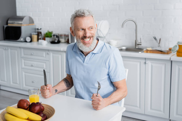 Smiling mature man holding cutlery near orange juice and fruits in kitchen  - 写真・画像