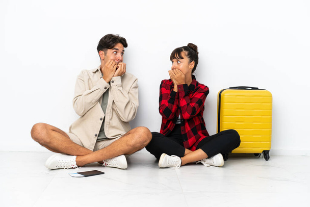 Jonge gemengde ras reizigers paar zitten op de vloer geïsoleerd op witte achtergrond is een beetje nerveus en bang het zetten van handen op de mond - Foto, afbeelding