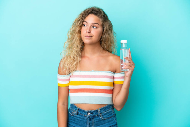 jong blond vrouw met een fles water geïsoleerd op blauwe achtergrond kijken naar de zijkant - Foto, afbeelding