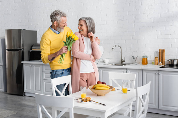 Cheerful man holding tulips near wife and breakfast in kitchen. Translation: "om, shanti, peace" - Photo, Image