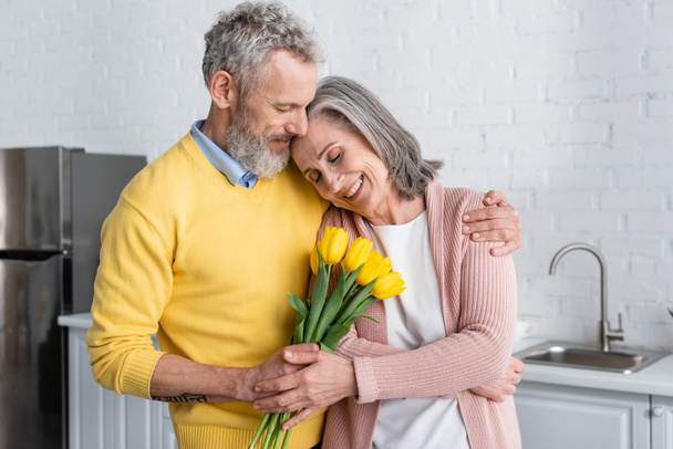 Middle aged man holding tulips near wife with closed eyes in kitchen  - Photo, Image