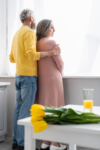 Homem abraçando esposa perto da janela e flores borradas na cozinha  - Foto, Imagem