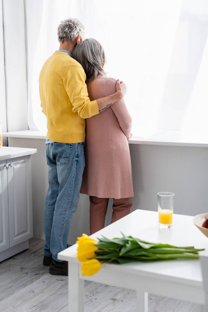 Back view of mature couple hugging near window and blurred tulips on table at home  - Foto, imagen