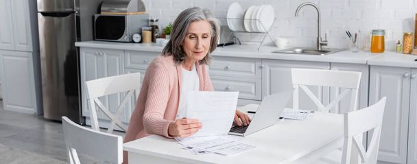 Siwe włosy dojrzała kobieta trzyma rachunki w pobliżu laptopa w kuchni, baner  - Zdjęcie, obraz