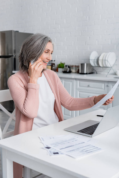 Smiling mature woman talking on smartphone and holding paper near laptop in kitchen  - Photo, Image