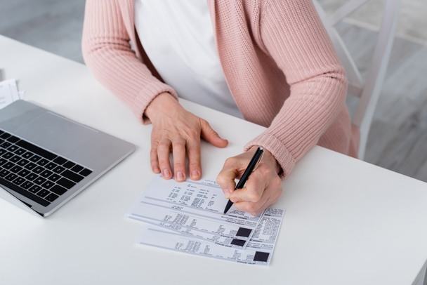 Καλλιεργημένη άποψη της γυναίκας που γράφει στο λογαριασμό κοντά στο φορητό υπολογιστή στο σπίτι  - Φωτογραφία, εικόνα