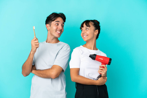 Jovem casal misto segurando um secador de cabelo e escova de dentes isolado no fundo azul olhando sobre o ombro com um sorriso - Foto, Imagem