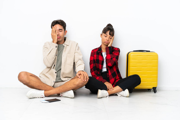 Jeune couple mixte de voyageurs assis sur le sol isolé sur fond blanc avec une expression faciale surprise et choquée - Photo, image