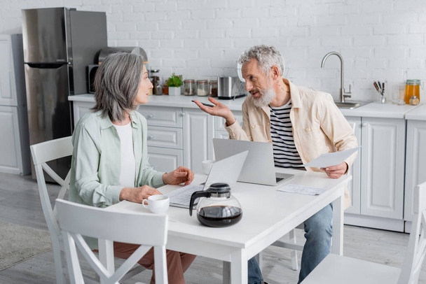 Mature couple talking near bills and laptops in kitchen  - Photo, Image