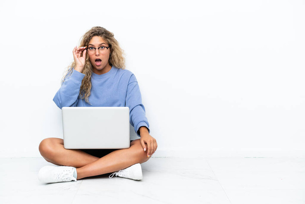 Meisje met krullend haar met een laptop zittend op de vloer met bril en verrast - Foto, afbeelding