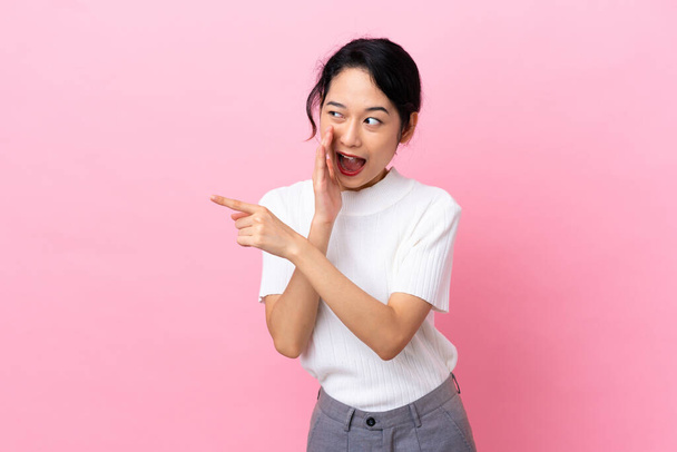 Young Vietnamese woman isolated on pink background pointing to the side to present a product and whispering something - Photo, Image