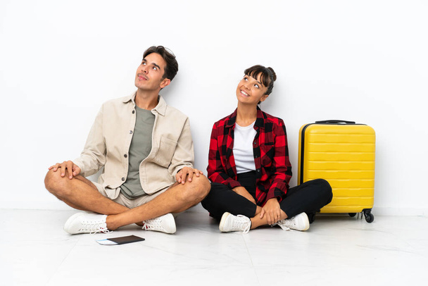 Mladý smíšený závod cestovatelé pár sedí na podlaze izolované na bílém pozadí pózování s pažemi v bok a smích - Fotografie, Obrázek