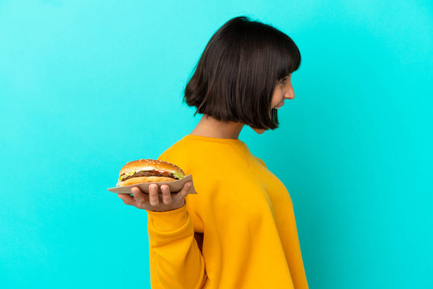 Junge brünette Frau hält einen Burger über isoliertem Hintergrund und lacht in seitlicher Position - Foto, Bild