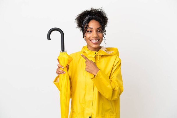 Mladá žena s nepromokavým pláštěm a deštníkem izolované na bílém pozadí ukazuje na stranu prezentovat produkt - Fotografie, Obrázek