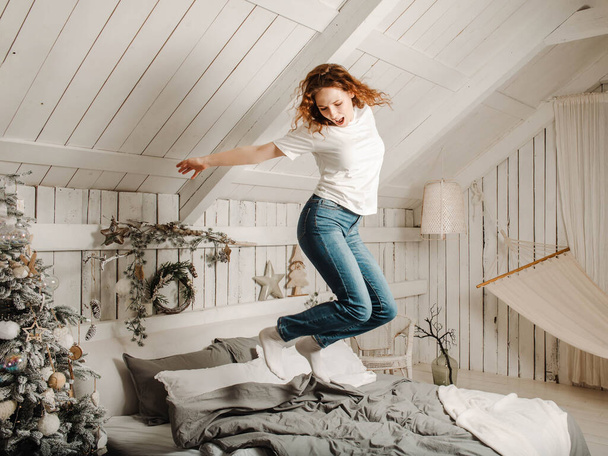 European young  fun beautiful woman with red hair in jeans jumps on a bed in a bright room with a Christmas interior, - Фото, зображення