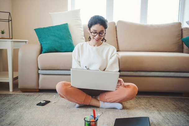 Front view of a woman, making notes while leaning on sofa and sitting on floor with laptop on crossed feet. Online education. Distance education. - Photo, Image