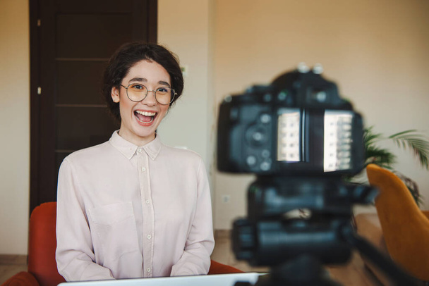 Καυκάσια δασκάλα καταγράφει μαθήματα βίντεο στην κάμερα χαμογελώντας και φορώντας γυαλιά. Διαδικτυακός τρόπος ζωής Εξ αποστάσεως εκπαίδευση. Βίντεο συνομιλία - Φωτογραφία, εικόνα