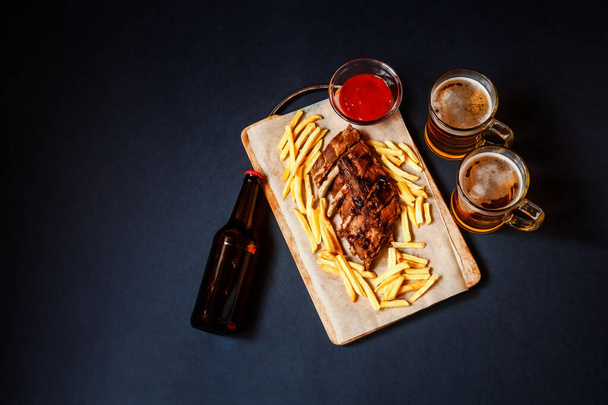 Baked pork ribs with french fries on cutting board and beer  on dark background. Top view, copy space. - Photo, Image