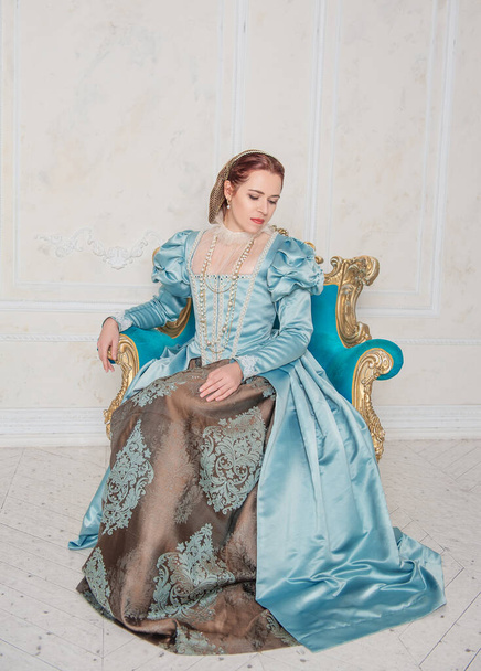 Beautiful young woman in medieval style blue dress sitting in the armchai - Photo, Image