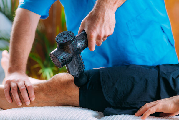 Terapeuta leczący mięśnie czworogłowe sportowca, używając pistoletu do masażu - Zdjęcie, obraz