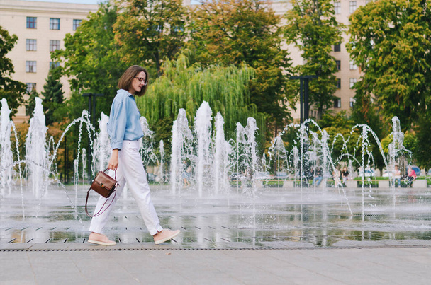 Urban portrait of young elegant business woman in eye glasses and casual clothes in city. Woman walking in green park, relaxing, smiling in sunny day. Hobby and leisure. - 写真・画像