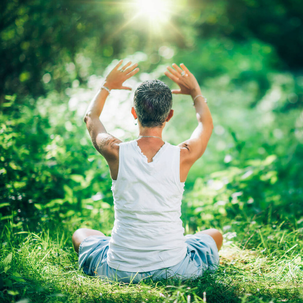Медитация для баланса и центрирования. Неузнаваемая внимательная женщина медитирует в пышном зеленом лесу, практикуя технику медитации, чтобы помочь своему центру и найти свой внутренний баланс. Сидя на земле, руки подняты над головой - Фото, изображение