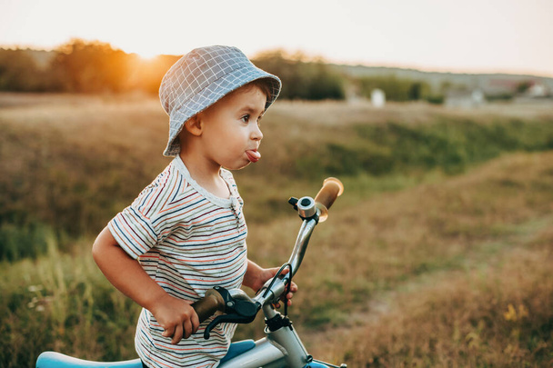 Portret chłopca jeżdżącego na rowerze po wiejskiej drodze spoglądającego z jednej strony w niebieskim kapeluszu i pokazującego język. Dzieciństwo - Zdjęcie, obraz