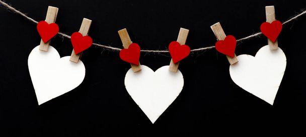 Happy Valentine 's Day Hintergrund Banner - Weißes Papier Notiz hängen auf hölzernen Wäscheklammern mit Holzherzen an einer Schnur isoliert auf dunkelschwarzer Textur, mit Platz für Text - Foto, Bild