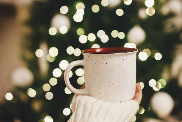 Hand in Hand im Pullover hält stilvolle Tasse auf dem Hintergrund des Weihnachtsbaums in Lichtern im Abendzimmer. Raum für Text. Keramische Tasse mit Tee in weiblicher Hand in festlichem Ambiente. Gemütliche Winterzeit - Foto, Bild
