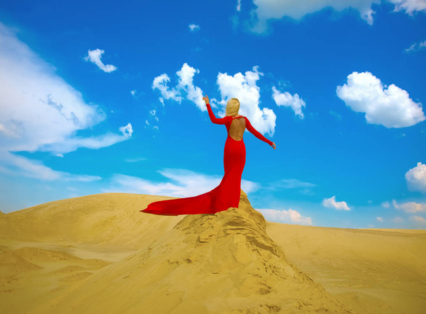 blonde woman posing in long red dress and  stand on sand desert against blue cloudy sky.  adult girl enjoying warm wind. Idea of of dry skin, Lack of water in body, outgoing time and youth. Back view - Foto, imagen