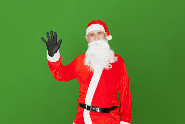 Un uomo caucasico vestito da Babbo Natale sta agitando con la mano aperta su cui indossa un guanto nero. Lo sfondo è verde. - Foto, immagini