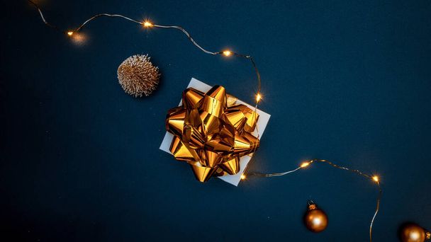 Christmas gift. White gift with golden bow, gold balls and sparkling lights garland in xmas decoration on dark background for greeting card. Xmas backdrop with space for text - Photo, Image