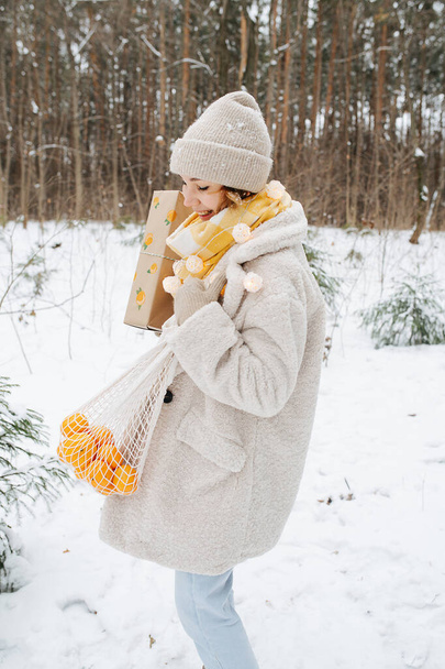 Cute cheerful young woman standing in a winter forest with present box and a net bag with tangerines. She's spinning in joy on Christmas Eve - Foto, Bild