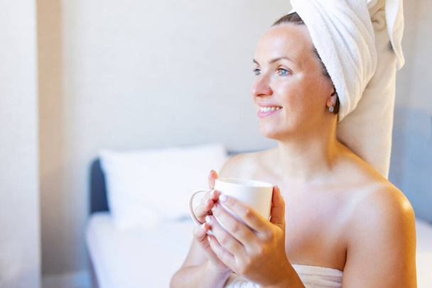 Attraktive junge Frau in Badetuch und Handtuch auf dem Haar hält eine Tasse und lächelt, während sie auf einem Bett in der Nähe des großen Fensters zu Hause sitzt. - Foto, Bild