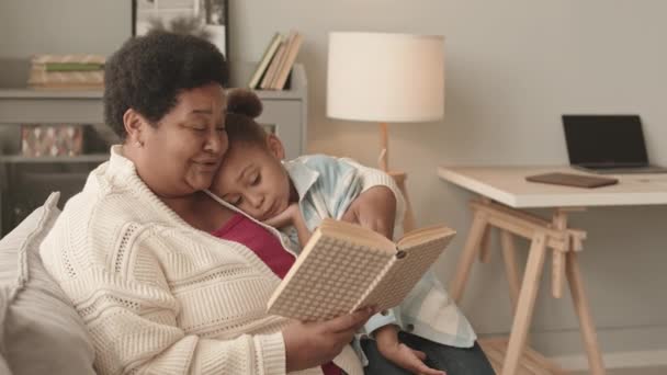 Keskipitkä iloinen viisivuotias tyttö istuu polvillaan hänen suosikki Afrikkalainen amerikkalainen isoäiti, joka lukee kirjaa ääneen olohuoneessa päivällä - Materiaali, video