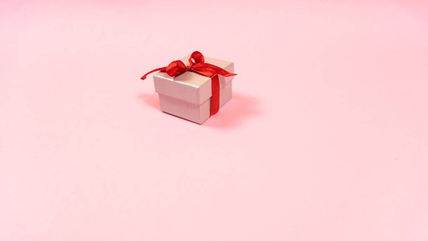 Petite boîte cadeau grise attachée avec un ruban rouge avec un cadeau pour un être cher isolé fond pastel rose clair. Espace de copie. Concept de préparation des cadeaux pour les vacances. - Photo, image