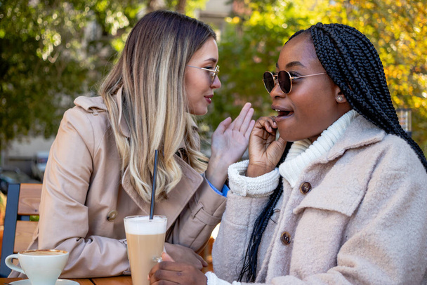 Two young girlfriends enjoy coffee together, sitting at a table, chatting, whispering, and gossiping. Diversity and leisure time concept. - Photo, Image